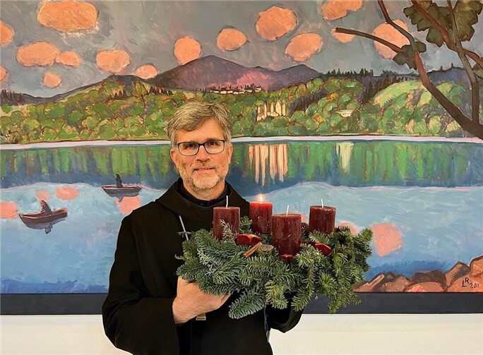 Abt Mauritius Wilde (OSB) mit einem Adventskranz – vor einem Gemälde, das den Laacher See und die Benediktinerabtei Maria Laach zeigt. Hier hat Mauritius Wilde im Oktober 2025 das Amt des Abtes übernommen. Das Gemälde im Hintergrund stammt von Künstlerbruder Lukas Ruegenberg („LR“) – einem 97jährigen Ordensmann, der in Maria Laach lebt. Foto: Robert Eberle / Maria Laach