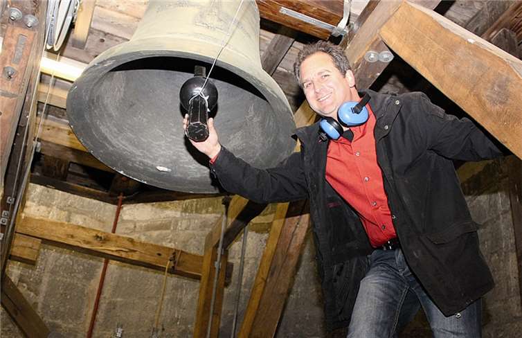 Andreas Bergmann ist mit einer kleinen Leiter weiter in den Glockenstuhl hinaufgestiegen und befestigt ein Seil an einem Glockenklöppel. BÜN