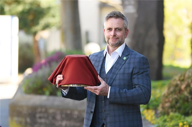 Bereits jetzt freut sich Landrat Marko Boos, den MYK-Pokal zum ersten Mal an eine Gemeinde oder Stadt im Landkreis Mayen-Koblenz überreichen zu können. Wie der neue Preis genau aussieht, bleibt bis zur Verleihung allerdings noch ein gut gehütetes Geheimnis. Foto: Foto: Kreisverwaltung MYK/teilweise KI-generiert