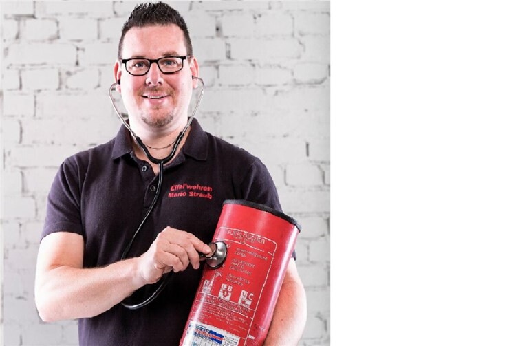 Bildunterschrift:Mit einem Augenzwinkern für mehr Sicherheit: Mario Straub von den Eifel´wehren „hört“ einen Feuerlöscher mit dem Stethoskop ab. Die humorvolle Aktion soll auf die regelmäßige Prüfung von Feuerlöschern nach DIN 14406-4 aufmerksam machen.