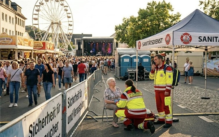 Das Bild zeigt eine belebte Kirmes oder ein Stadtfest mit einem Riesenrad, Ständen und einer Konzertbühne im Hintergrund. Im Vordergrund kümmern sich Einsatzkräfte eines Sanitätsdienstes an einem Zelt um eine ältere Frau, die auf einem Stuhl sitzt.