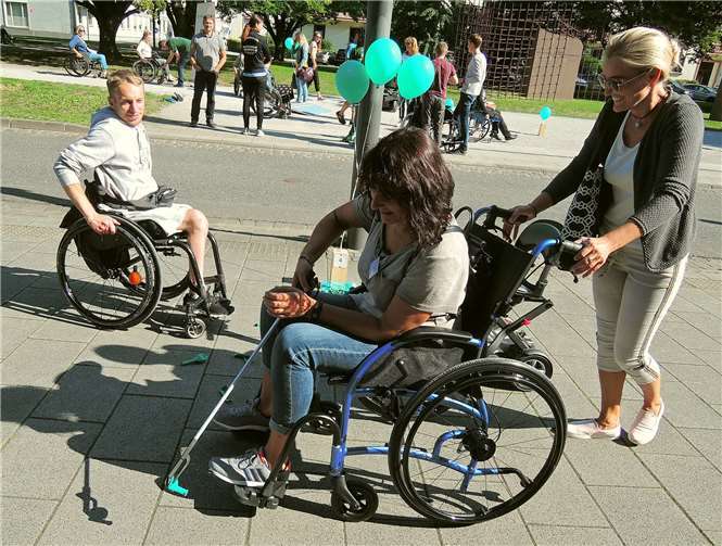 Der junge Mann im Hintergrund, der seit frühester Kindheit an den Rollstuhl gefesselt ist, beobachtet beinahe amüsiert, wie die nicht behinderte Dame an die Greifhilfen-Aufgabe herangeht.