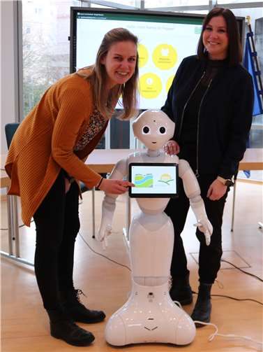 Die Digitalisierungsbeauftragte der Verbandsgemeinde (VG) Weißenthurm, Lena Rasch (li.) und Alessa Strubel, Projektmanagerin von „Starke Kommunen - Starkes Land“ nehmen Pepper zum Start des Pilotprojektes in der VG Weißenthurm in Empfang. Foto: VG Weißenthurm