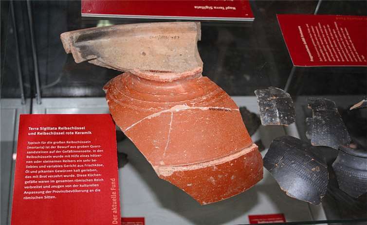 Die rötlichen Fragmente stammen von einer Reibschüssel und zeigen an der Innenseite den typischen Bewurf grober Quarzpartikel. Die schwarz glänzenden Scherben gehören zu dünnwandigen Schank- und Trinkgefäßen.