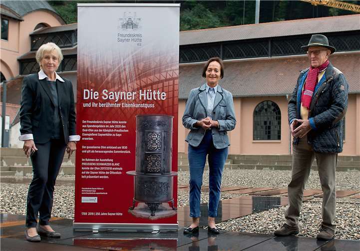 Freundeskreis-Vorsitzende Rehlinde Glöckner bedankt sich zusammen mit Vorstandsmitglied Werner Hennemann bei Johanna Bernardi für die Spende, die dem Freundeskreis den Erwerb des Eisenguss-Ofens vom Deutschen Ofen-Museum ermöglichte. Das Banner zeigt eine Abbildung des Oval-Ofens, der künftig wichtiges Exponat der Museumsausstellung sein wird. Foto: Freundeskreis Sayner Hütte e.V./Benedikt Rothkegel