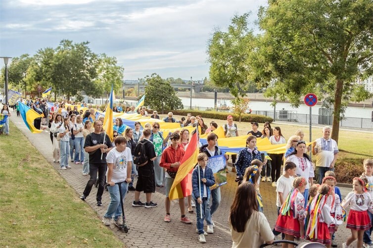 Kinder in ukrainischen Trachten, Familien und Unterstützer marschieren gemeinsam am Rheinufer — auch eine deutsche Fahne ist dabei.