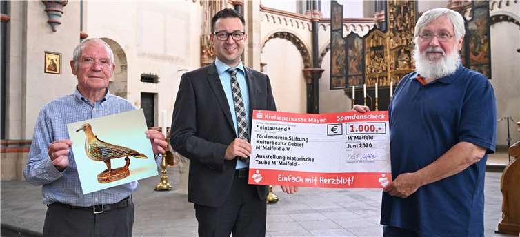 Martin Steinsiek als Leiter des BeratungsCenters der KSK in Münstermaifeld (Mitte) gratuliert den beiden Vorstandsmitgliedern Dieter Ritzenhofen (links) und Otto Wölbert (rechts) persönlich zur Spende und übergibt symbolisch den „großen“ Scheck im Namen der Kreissparkasse Mayen. Auf dem Bild links ist eine Abbildung der Münstermaifelder Eucharistietaube zu sehen. Foto: privat