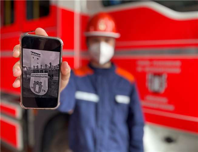 Symbolbild im Jubiläumsjahr: Jugendfeuerwehrmitglied Robin, geschützt mit einer FFP2-Maske, und seinem Smartphone. Auf diesem zu sehen die Gründungsmitglieder von 1976, die noch heute Mitglied der Weißenthurmer Feuerwehr sind. Foto: E. Brüning