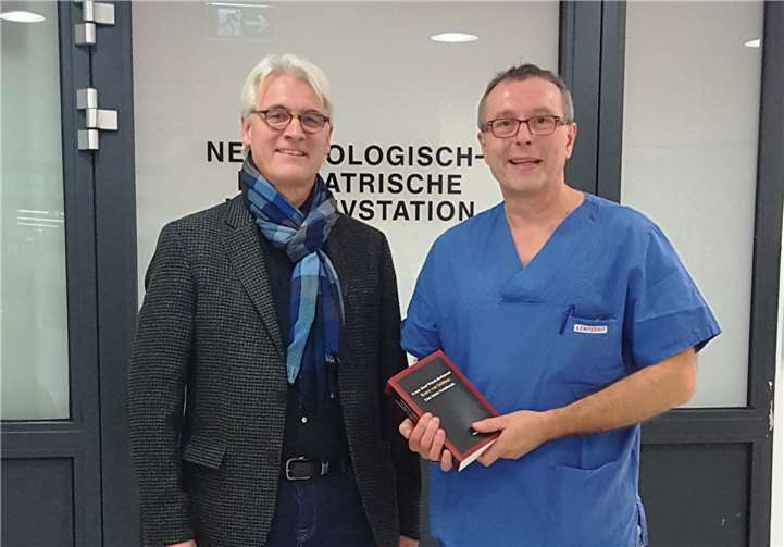 Vor dem Eingang zur Frühgeborenen-Intensivstation am Kemperhof: Dr. Franz Josef Witsch-Rothmund (li.) überreicht ein Exemplar seines Buches und die Spende über 1.000 Euro an Oberarzt Dr. Thomas Hoppen. privat