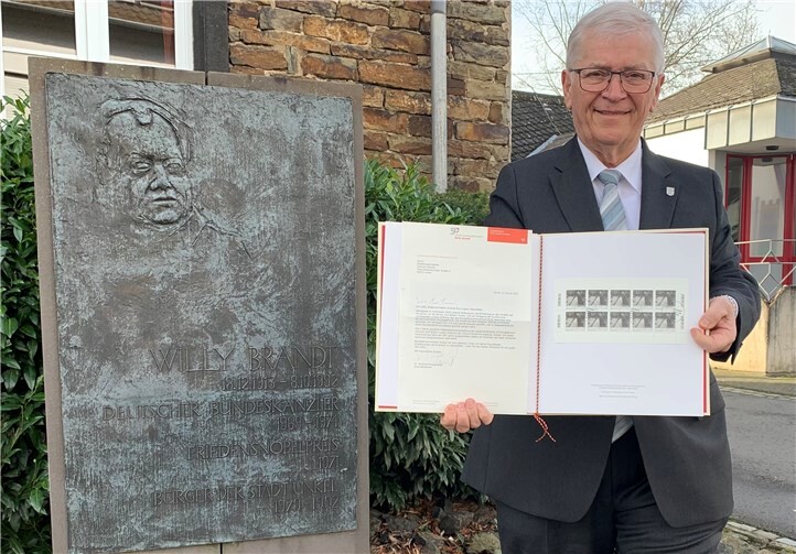 Vor der Gedenktafel des Unkeler Bürgers, Bundeskanzlers und Friedensnobelpreisträgers Willy Brandt vor dem Historischen Rathaus der Kulturstadt präsentiert Stadtbürgermeister Gerhard Hausen die stark limitierte Schmuckausgabe-Mappe mit der Sonderbriefmarke anlässlich des 50. Jahrestages des historischen Kniefalls von Warschau. Foto: Thomas Herschbach/Kulturstadt Unkel